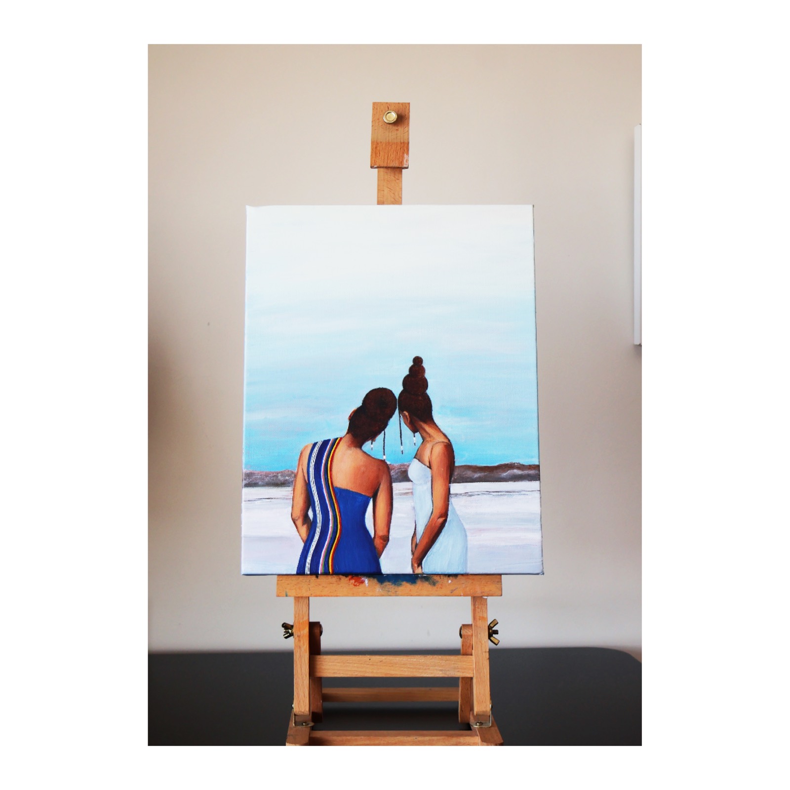 Two beneath a heavy sky — two women seen from behind on a pale salt pan, heads inclined toward the horizon, beaded strands falling between them; photographed on an easel